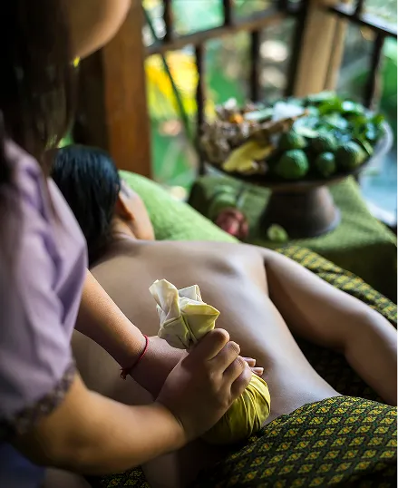 Woman receiving Ayurveda Pinda Massage on a green cushioned bed, with fruits and vegetables on a tray in front of her.
