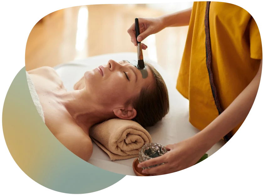 A woman receiving a massage in a spa setting, with a therapist applying a treatment solution to her forehead.