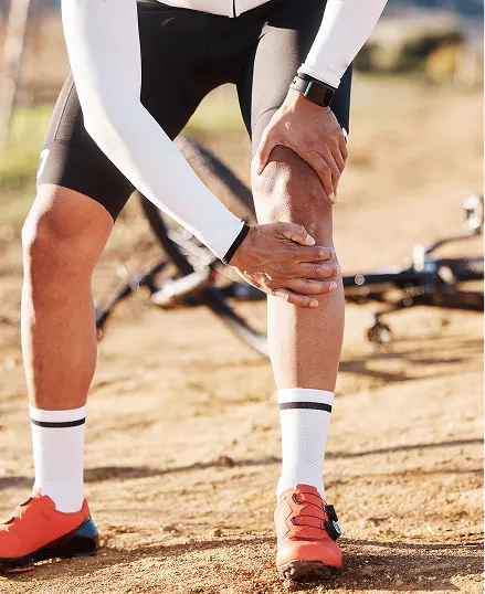 A person in a cycling stance with their leg bent, wearing a white and black cycling outfit, on a dirt path with a bicycle in the background.