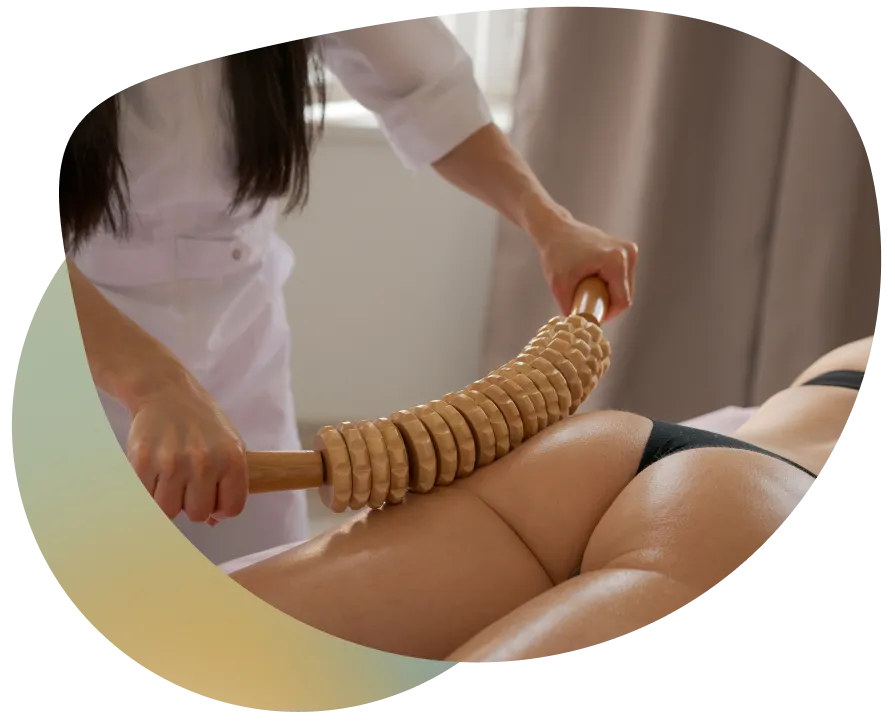 A woman is massaging her back with a wooden brush, lying on a bed with a window in the background.